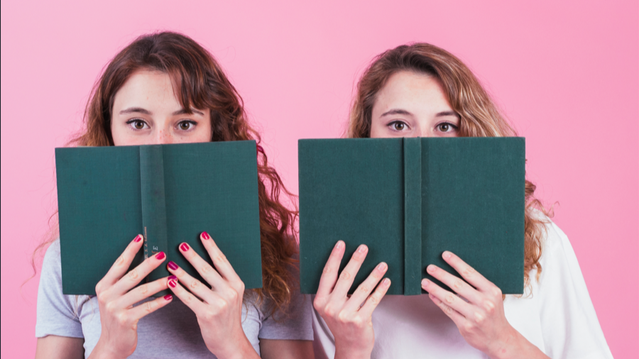 Dos amigas cubriendo su rostro con el libro de tapa verde sobre fondo rosa